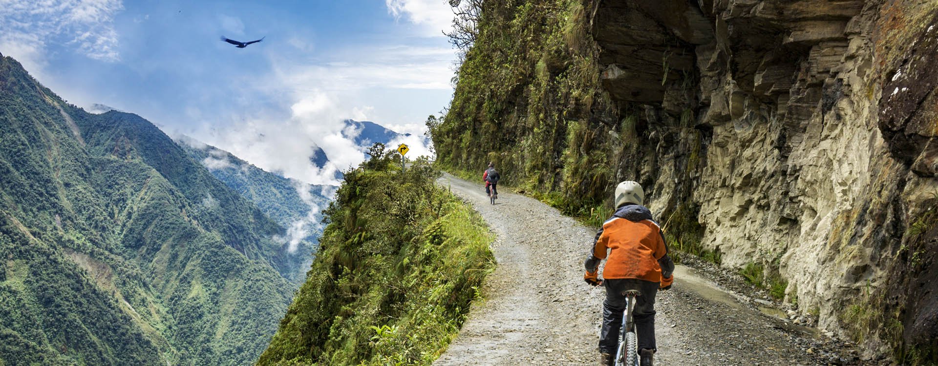 Bike adventure travel photo. Bike tourists ride on the "road of death" downhill track in Bolivia. In the background sky circles a condor over the scene.