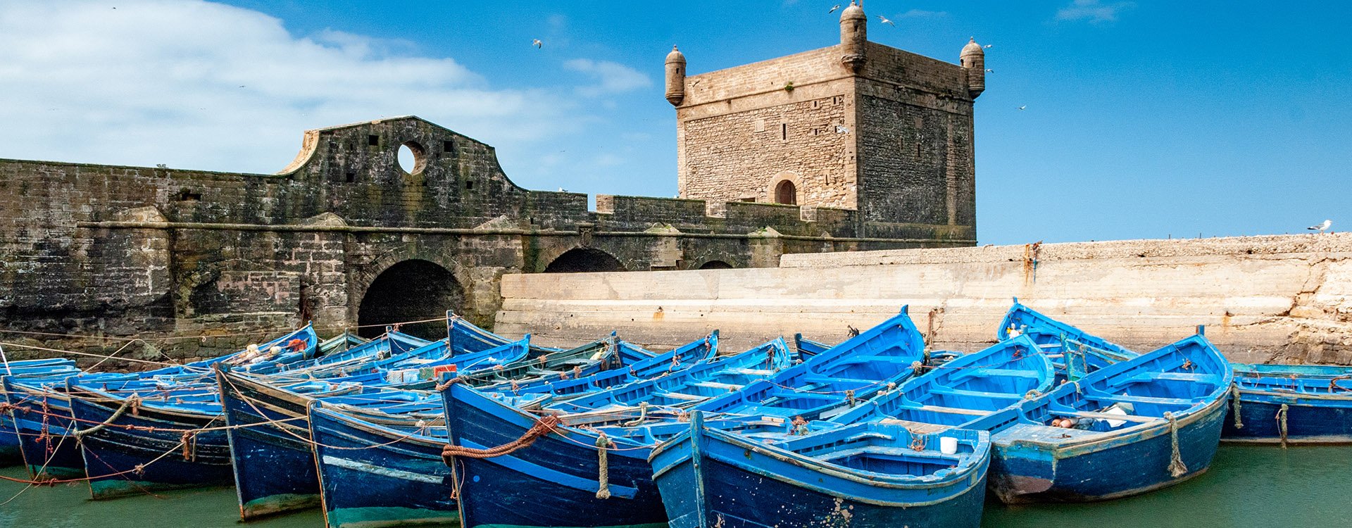 A fleet of blue fishing boats in the port of Essaouira in Morocco. Tower of the citadel of Mogador