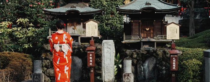 A woman dressed in a vibrant red kimono with seasonal patterns, stands respectfully in front of two small shrine buildings.