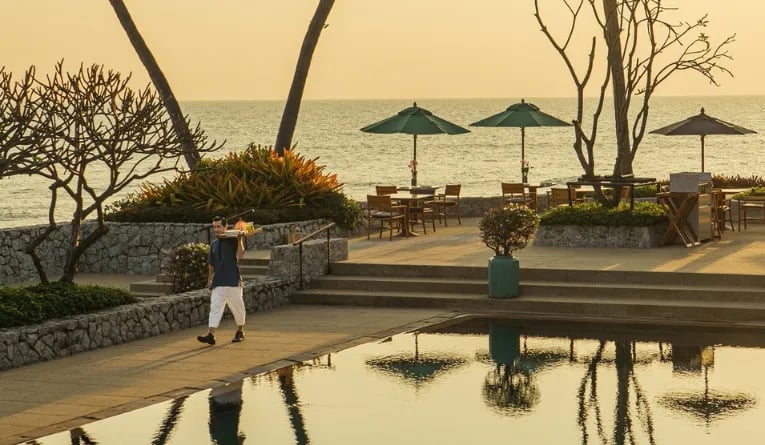 The pool pavillion at Chiva-Som in Hua Hin, Thailand, at sunset, with dining tables, green umbrellas, palm trees, and swimming pool overlooking the sea