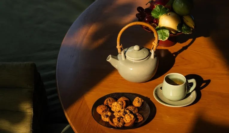 A teapot, a cup of tea, a plate of small baked snacks, and a bowl of fruit on a wooden table